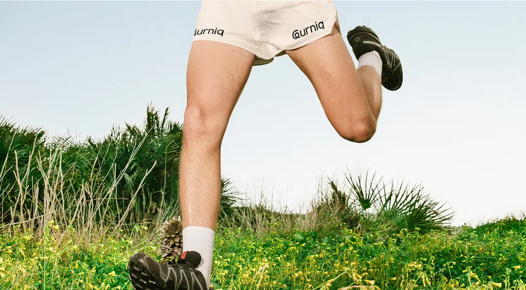 Leurniq wordmark on cream running shorts, mid-stride in a grassy field under a pale sky