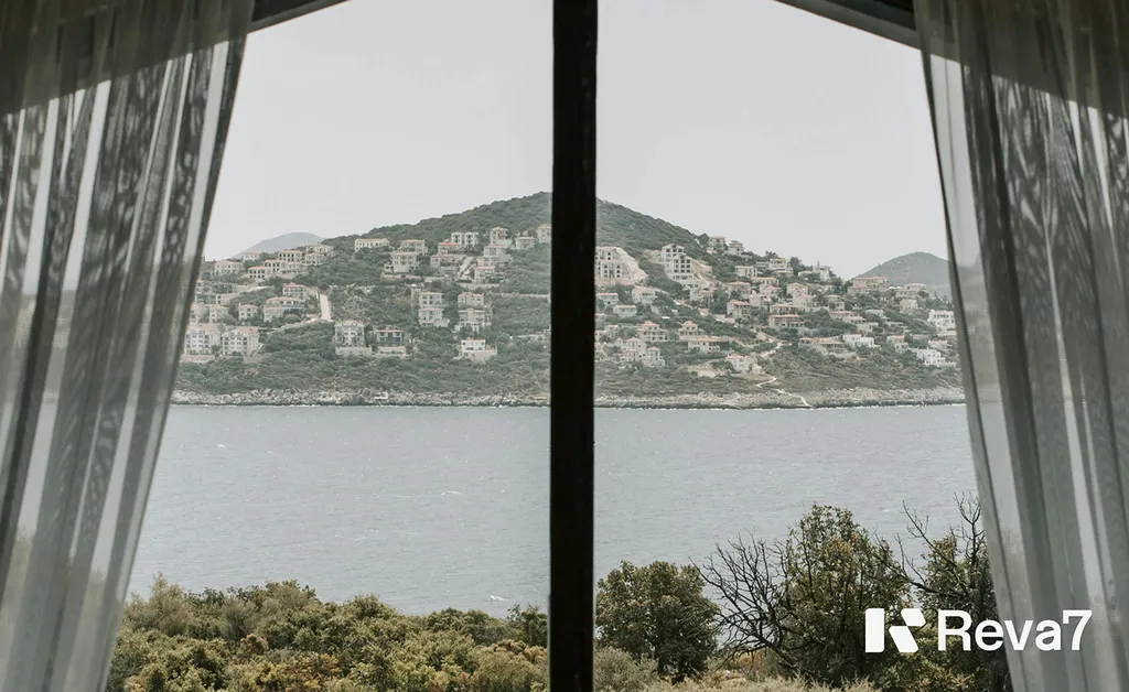 View from a curtained window across the sea to a hillside town, with the Reva7 logo